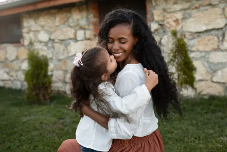 A smiling mother and her young daughter sharing a warm hug outdoors.