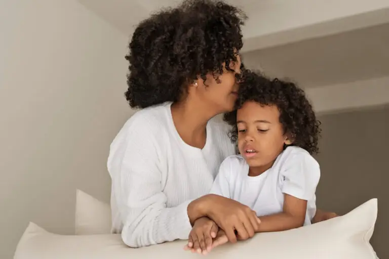 A mother affectionately kissing her young daughter's head indoors.